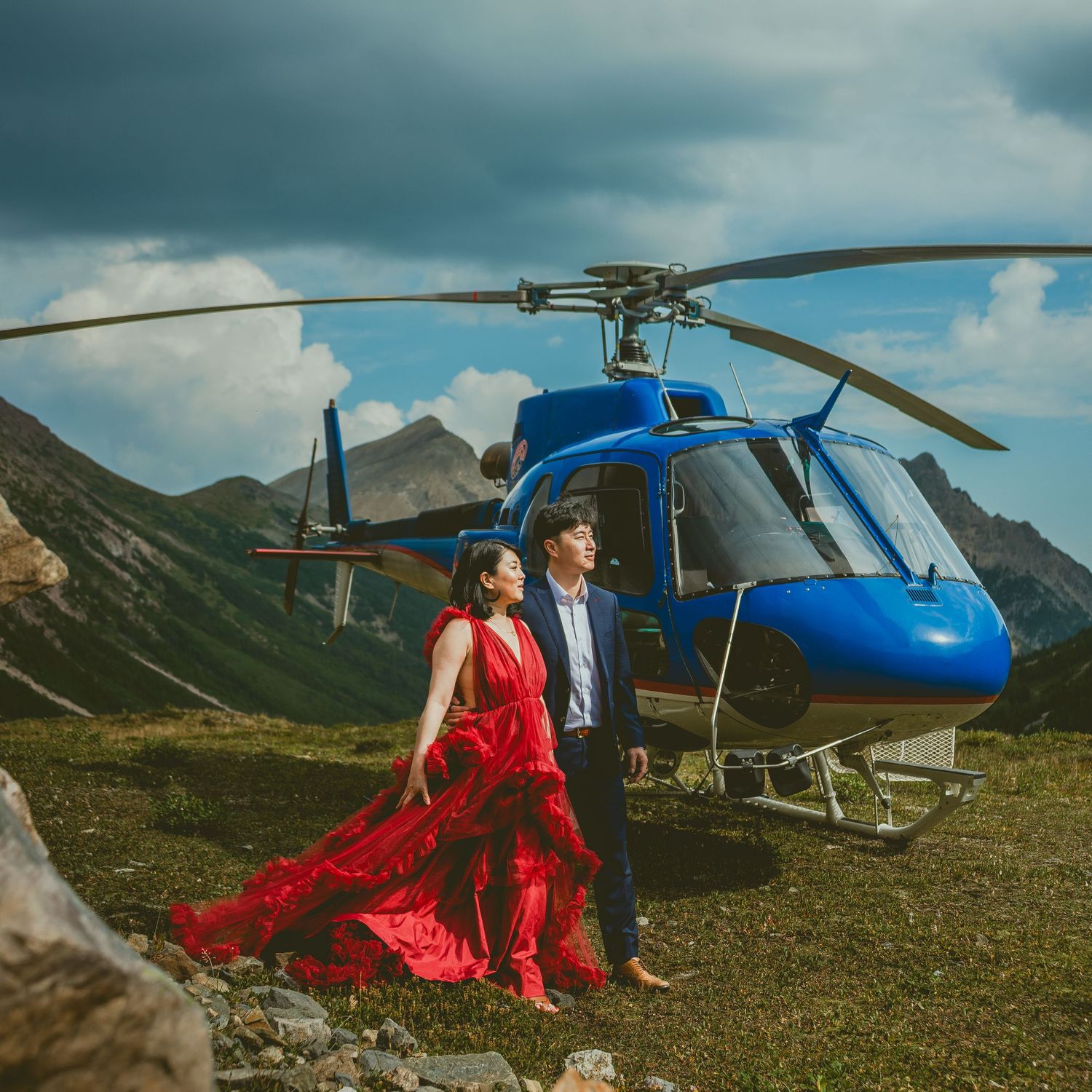 Helicopter parked in dramatic mountain landscape with moody storm clouds and bride in flowing red dress.