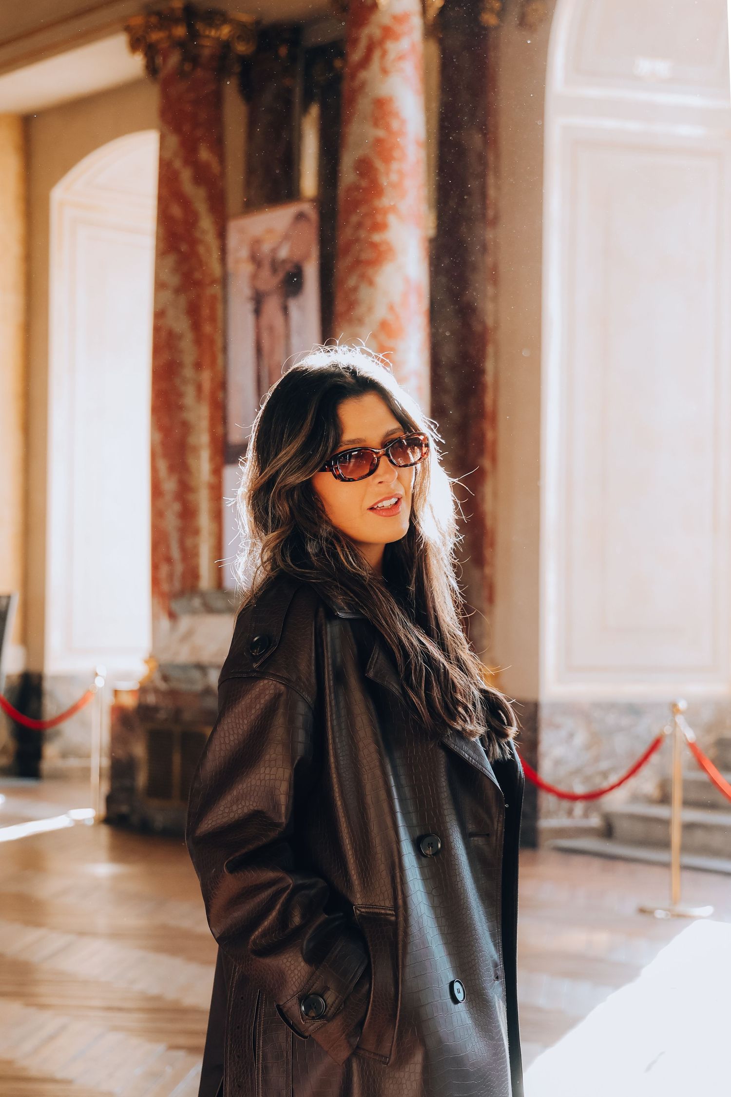 A stylish woman in a dark trench coat and brown sunglasses poses inside an ornate classical building.