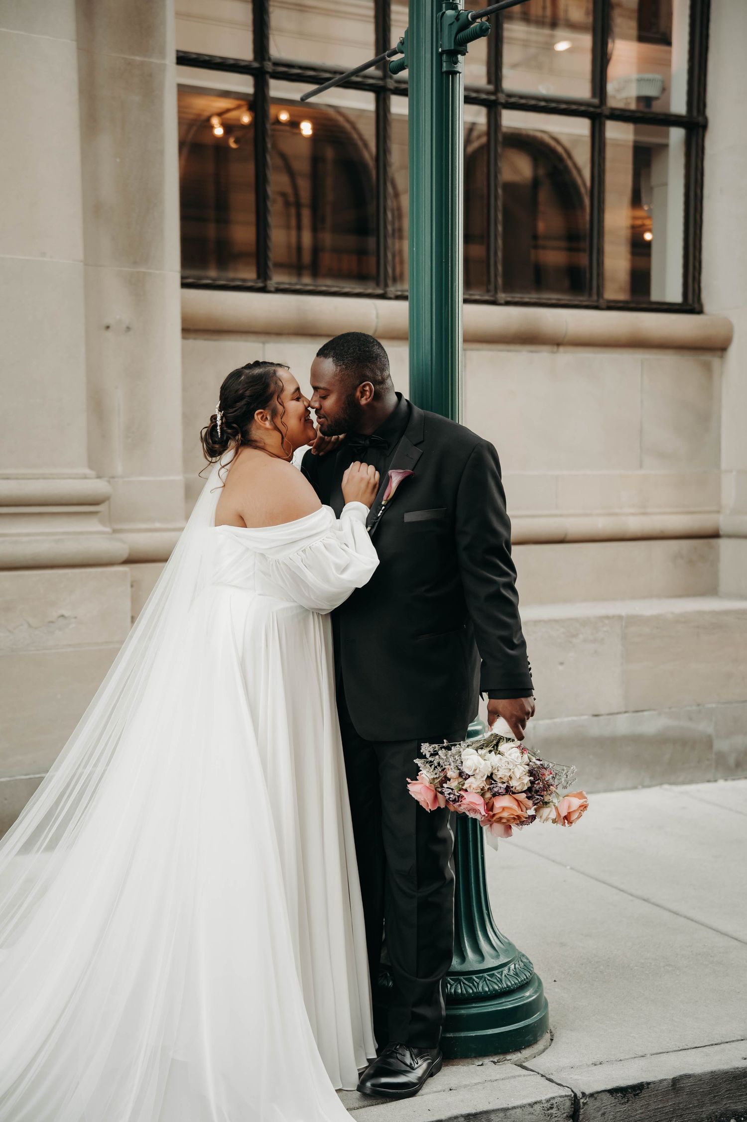 Bride in off-shoulder white gown and groom in black tuxedo share a kiss beside a green lamppost on a city street.