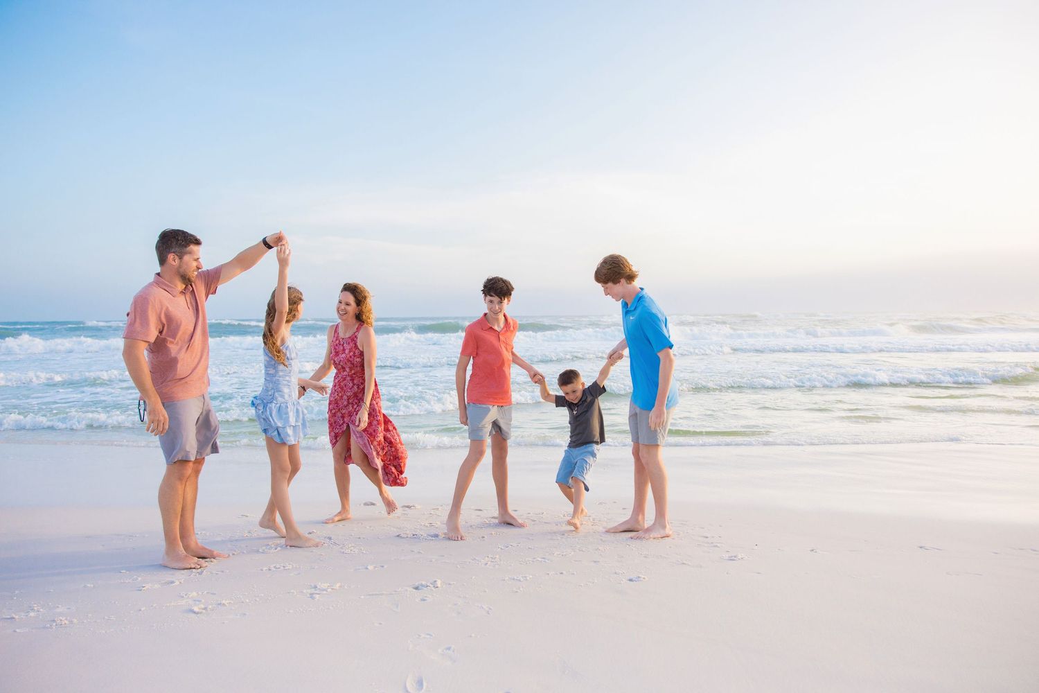 A family in light colored clothing explores the beach at sunset while collecting seashells along the shoreline.