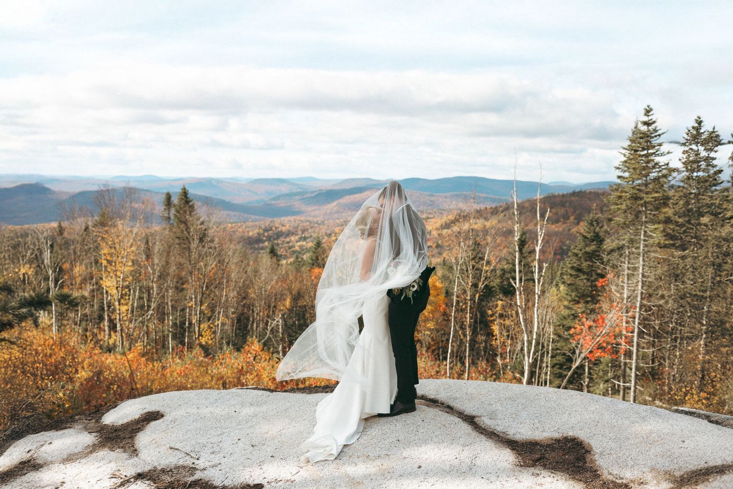 A couple in wedding attire stands on a snowy mountain peak overlooking autumn foliage and rolling hills in the distance.