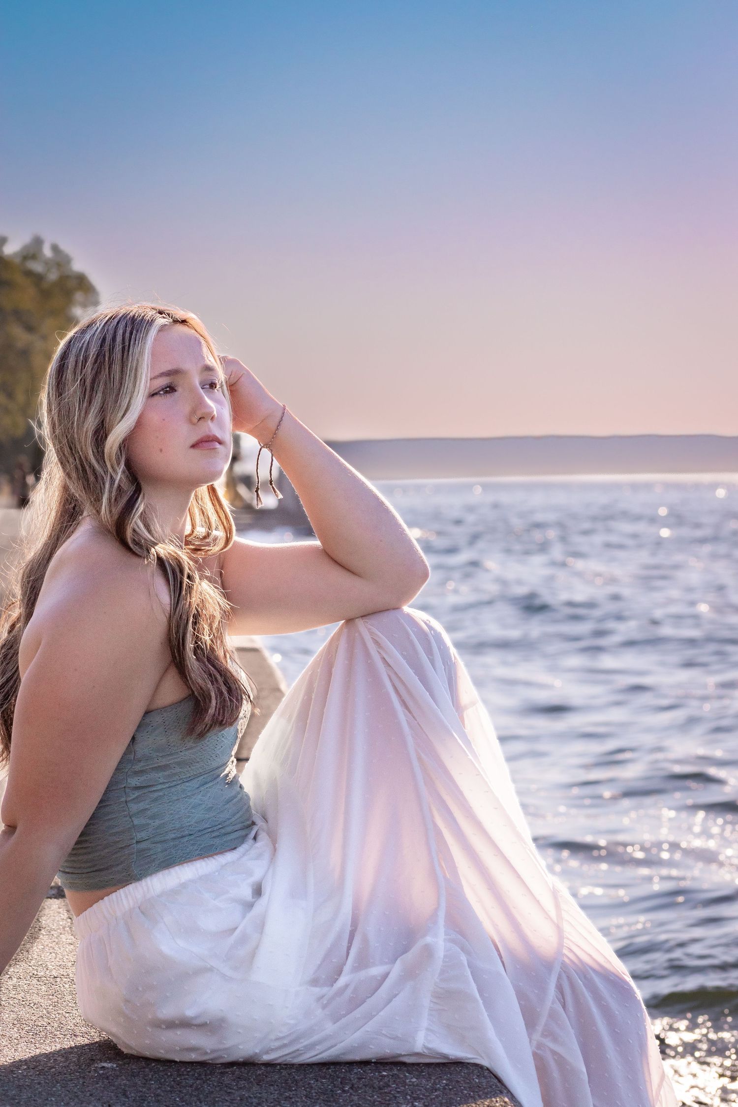 Senior girl in white skirt and sage green top sitting on the seawall at Alki Beach, with Seattle waterfront behind her.