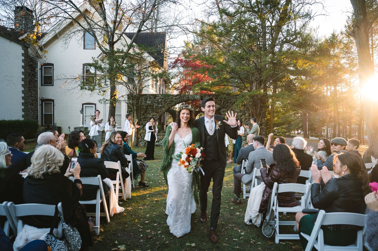 Hudson Valley, NY wedding couple exit outdoor ceremony up lawn aisle with fall foliage and stone arches at sunset at Brotherhood Winery.