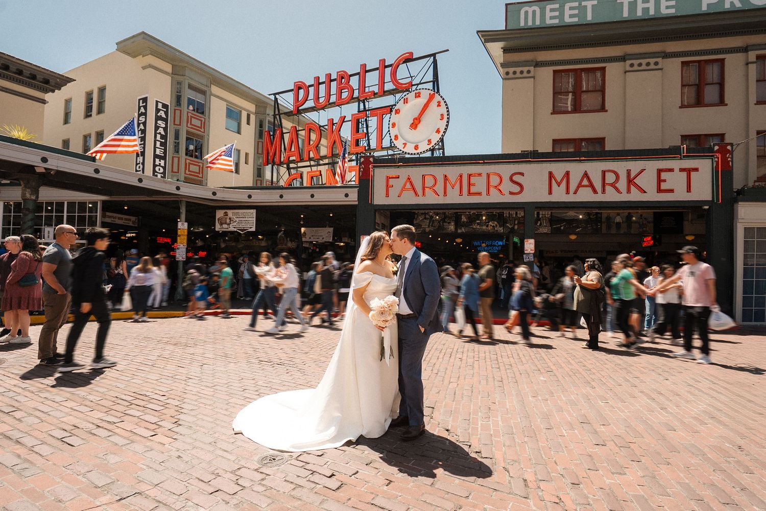 A couple shares a romantic moment in front of the iconic Pike Place Market sign in downtown Seattle on a sunny day.
