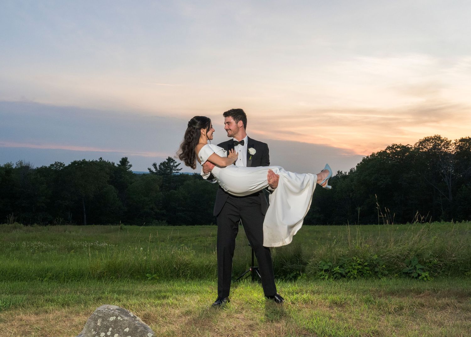 A couple shares a romantic moment at sunset, with the groom carrying the bride in a grassy field.