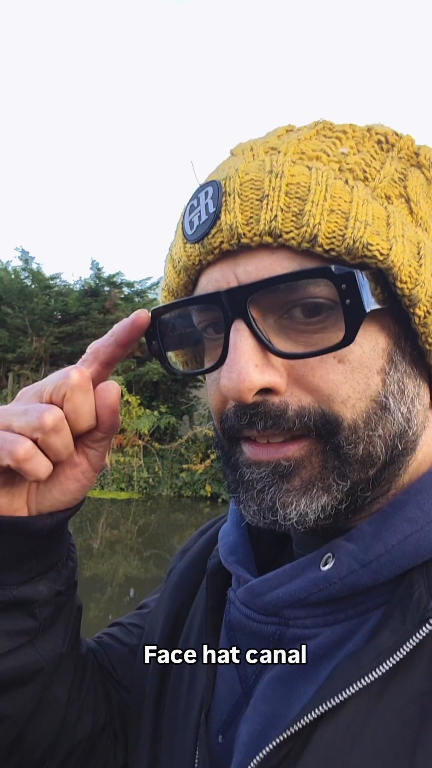 A person wearing a yellow knit hat and glasses smiles while taking selfies near a lake on a sunny day.