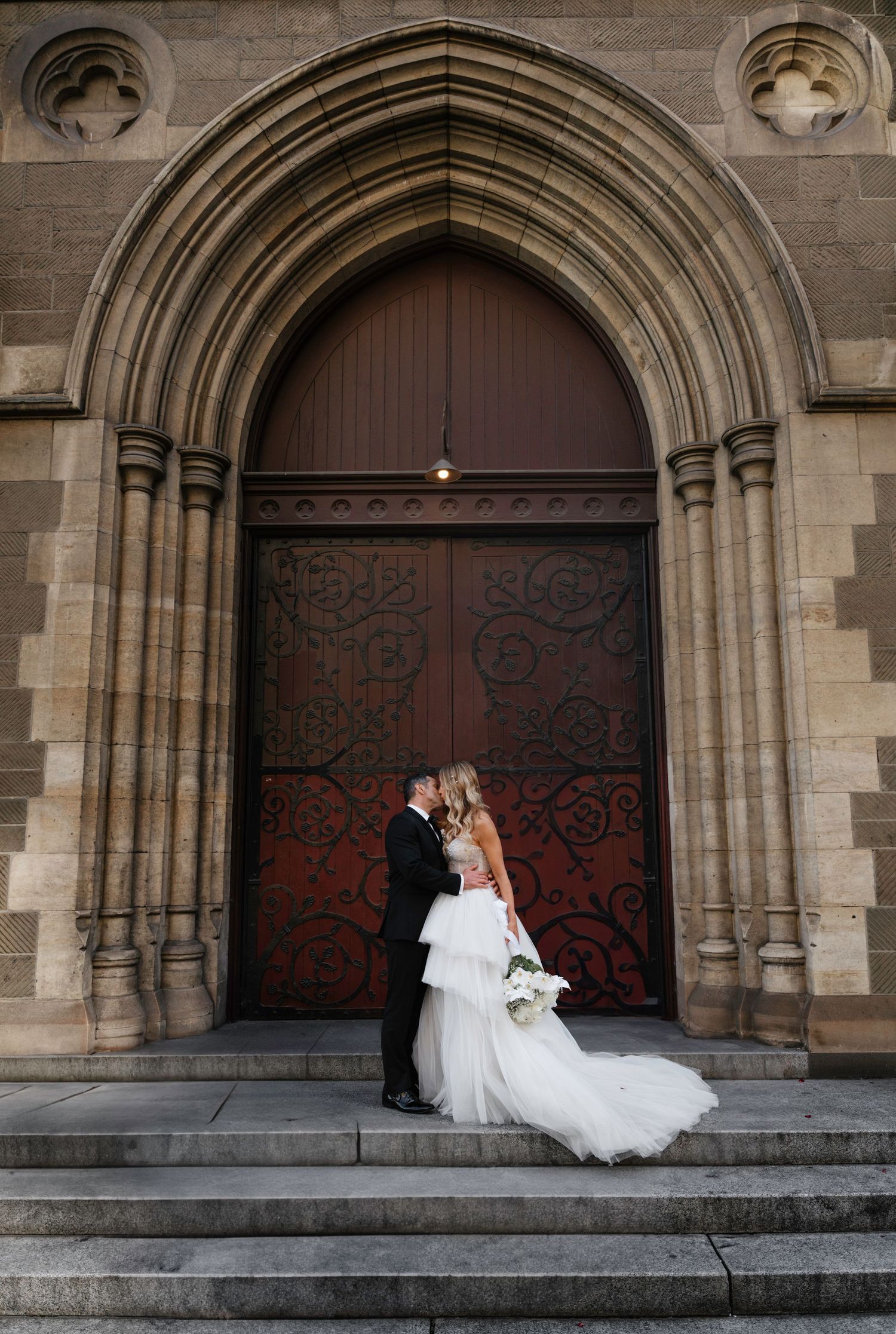 Elegant couple stands in front of ornate Gothic church doorway with grand arched entrance and decorative stonework.