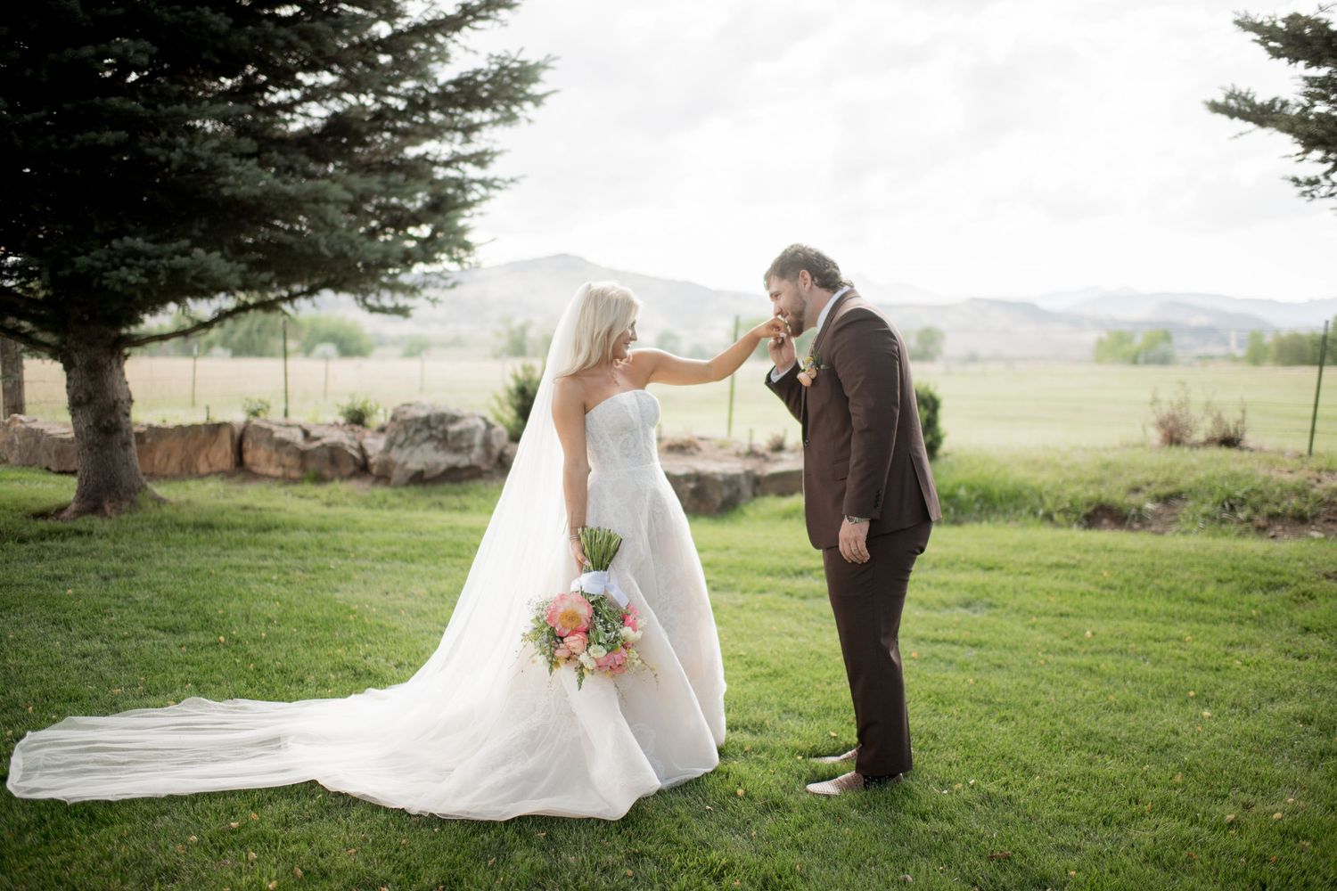 A wedding couple poses in a scenic green field with mountains and trees in the background during their outdoor ceremony.