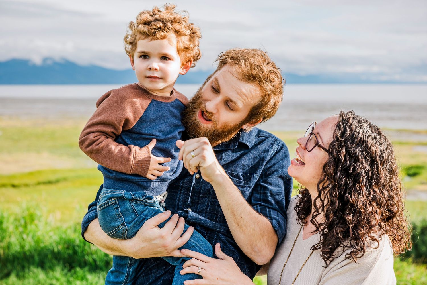 Family enjoying tender moments together outdoors in a grassy field with mountains in the background.