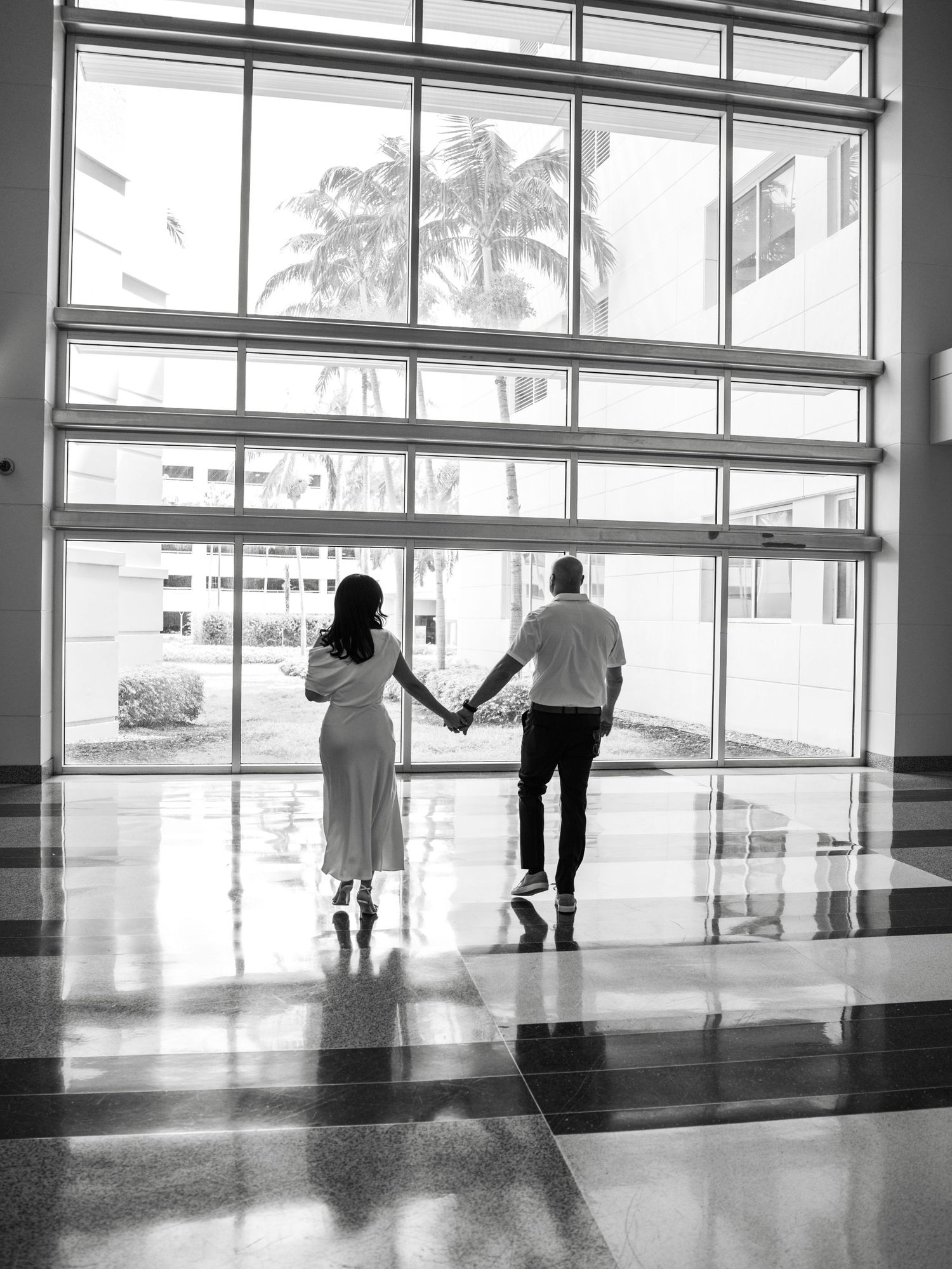 A couple holds hands while walking through a modern building with large windows and palm trees visible outside.