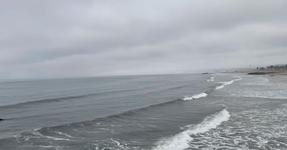 An aerial view of the California coastline shows waves crashing along a sandy beach on a cloudy day.