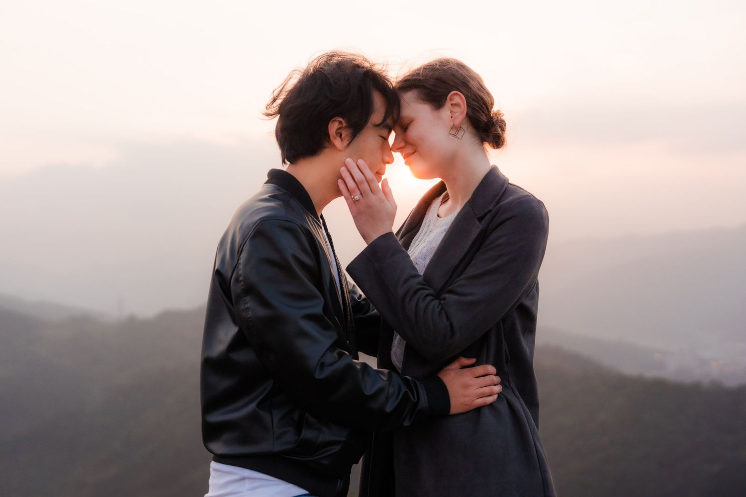 Two people share an intimate moment at sunset against a mountain landscape backdrop.