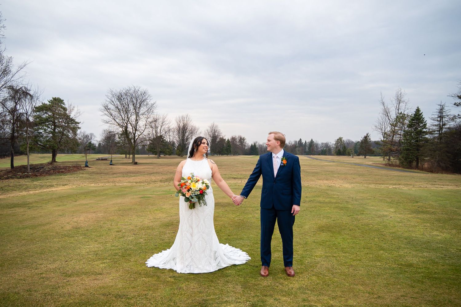 A couple in wedding attire walks hand in hand across a scenic open field on an overcast autumn day.