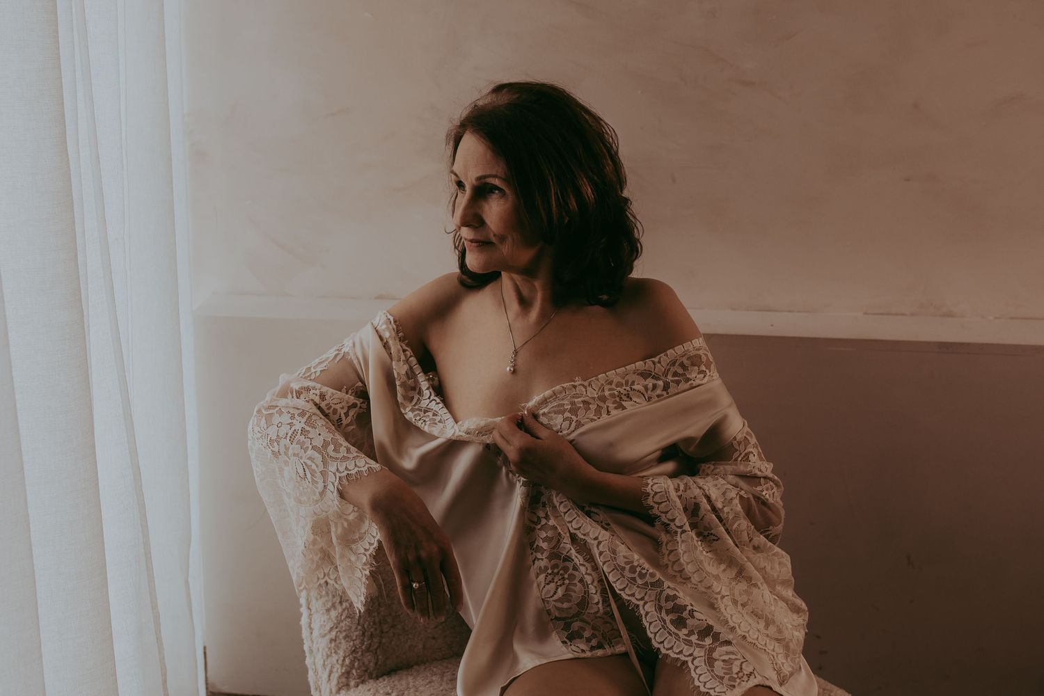 Oudere vrouw die een boudoirshoot heeft gehad waarbij ze een witte kimono aan heeft met kant op een stoel met een witte achtergrond