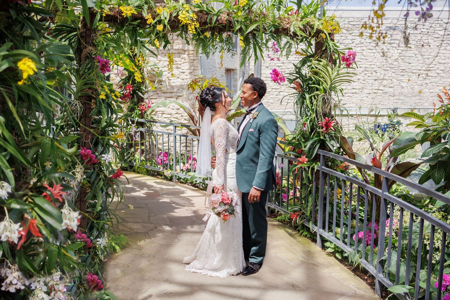 A bride and groom poses together in a romantic Dallas garden wedding venue decorated with pink and red flowers and greenery along a railing.