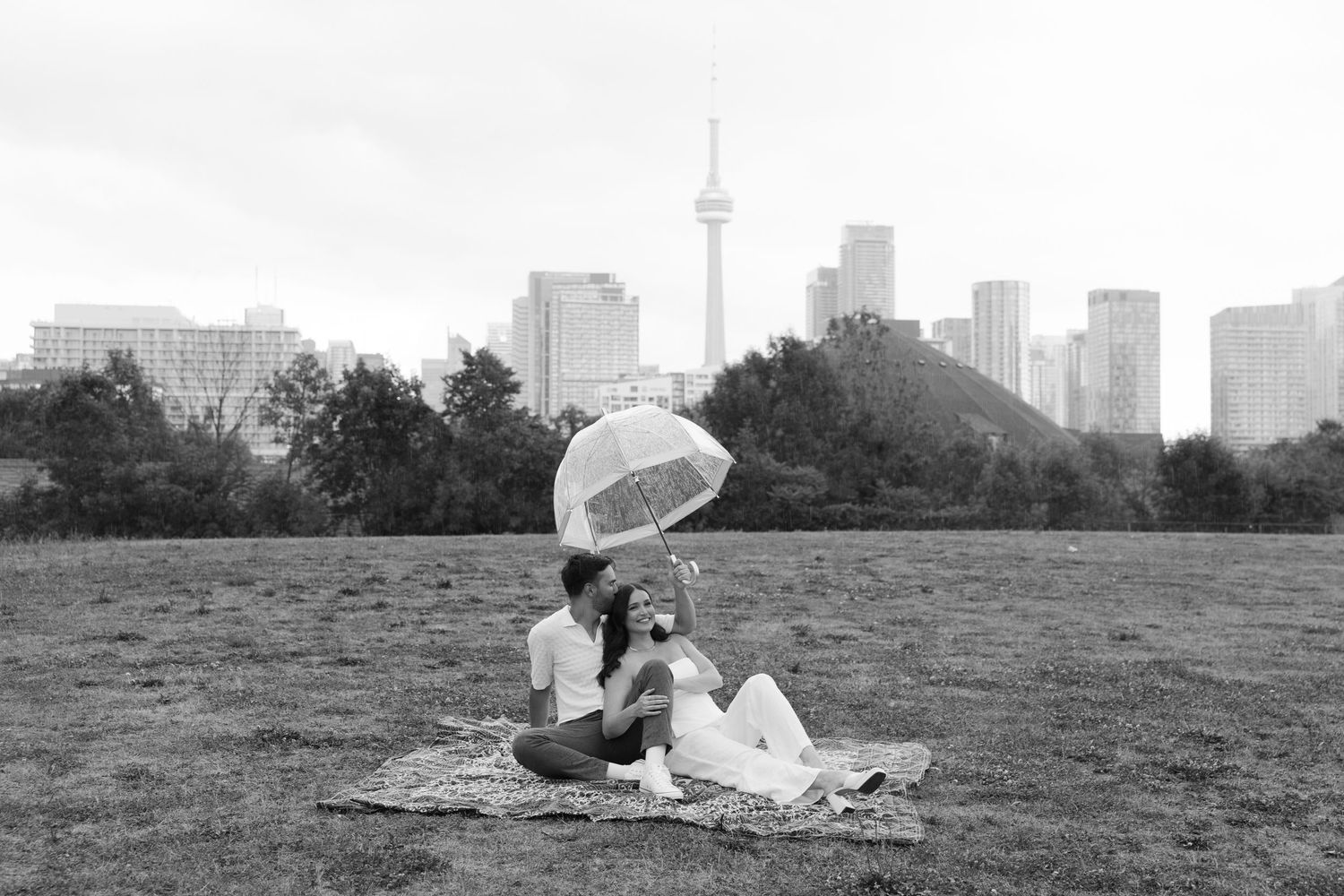 A couple enjoys a romantic picnic on a blanket with clear umbrella and Toronto skyline featuring CN Tower in background.