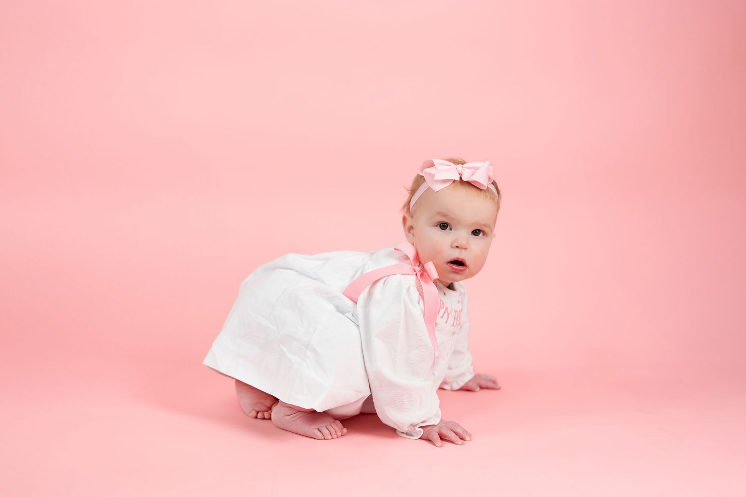 Adorable baby girl in white dress and pink bow headband crawling on pink studio background.