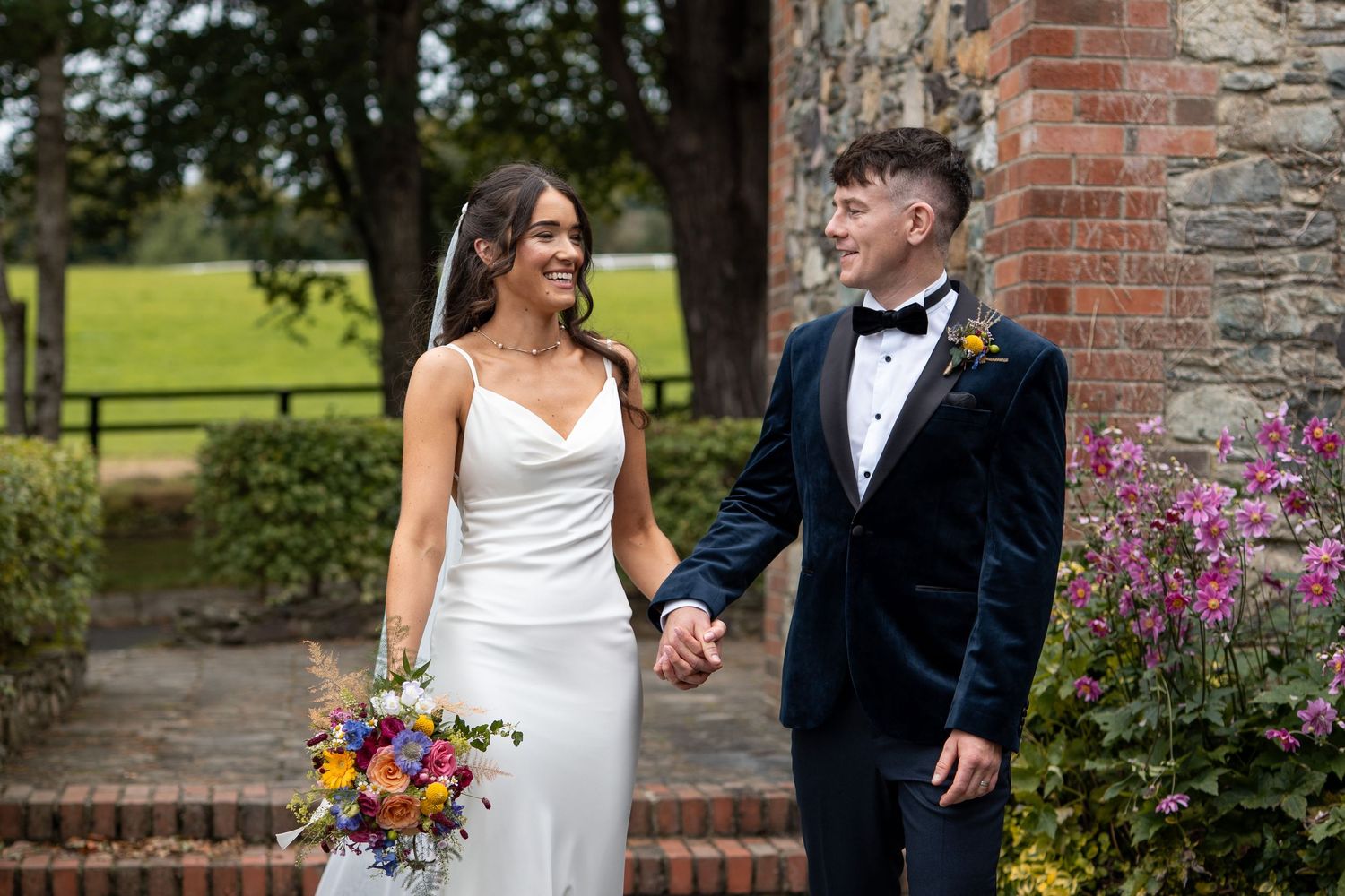 A couple in wedding attire walks and laughs together in a garden setting with brick walls and colorful flowers.