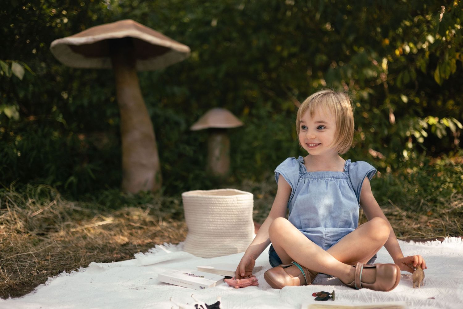 A little girl sits on a blanket with toys in front of a few giant mushrooms.