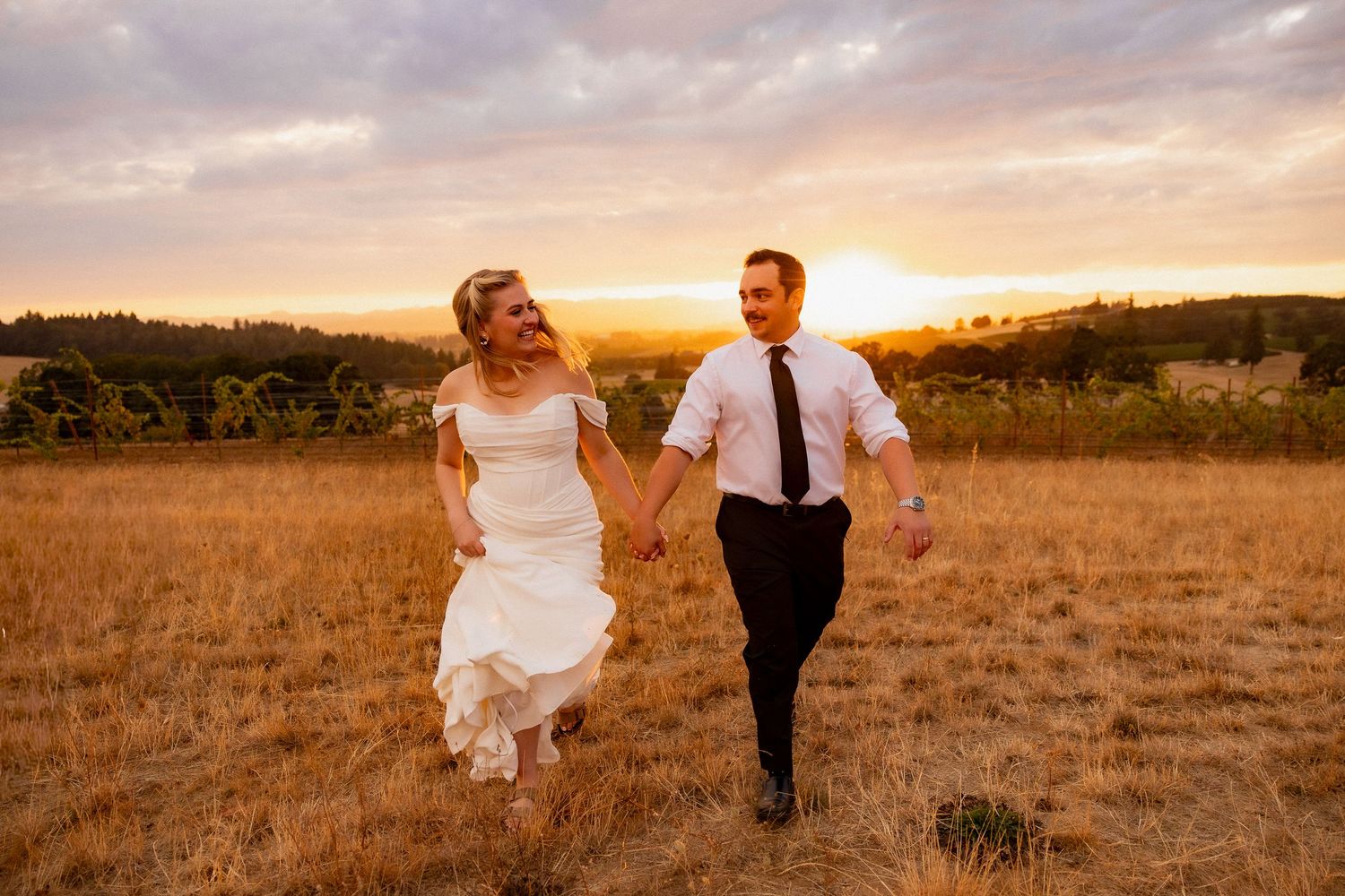 A couple in elegant wedding attire stand together outside a modern building at dusk with warm lighting.