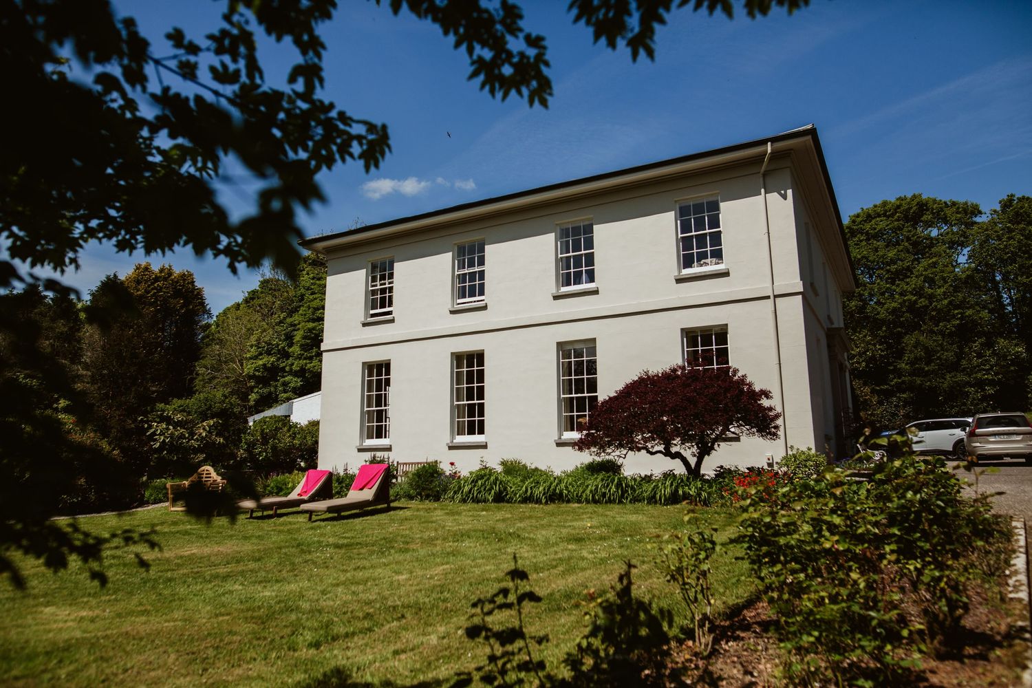 White two-story Georgian style house with symmetrical windows and red lounge chairs on the lawn on a sunny day.