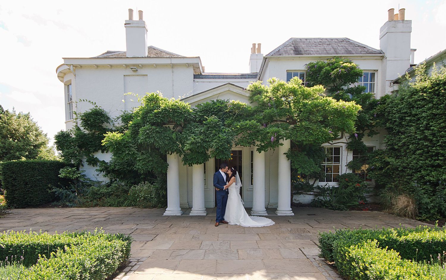 A couple walks through a scenic park with trees and a winding stream on their wedding day.