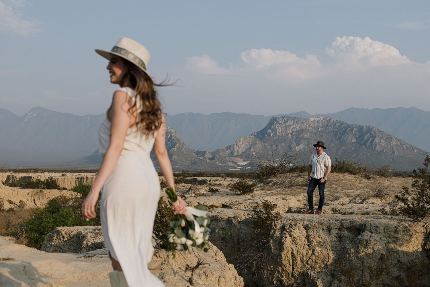 Bride in flowing dress stands on rocky terrain with mountains in background.