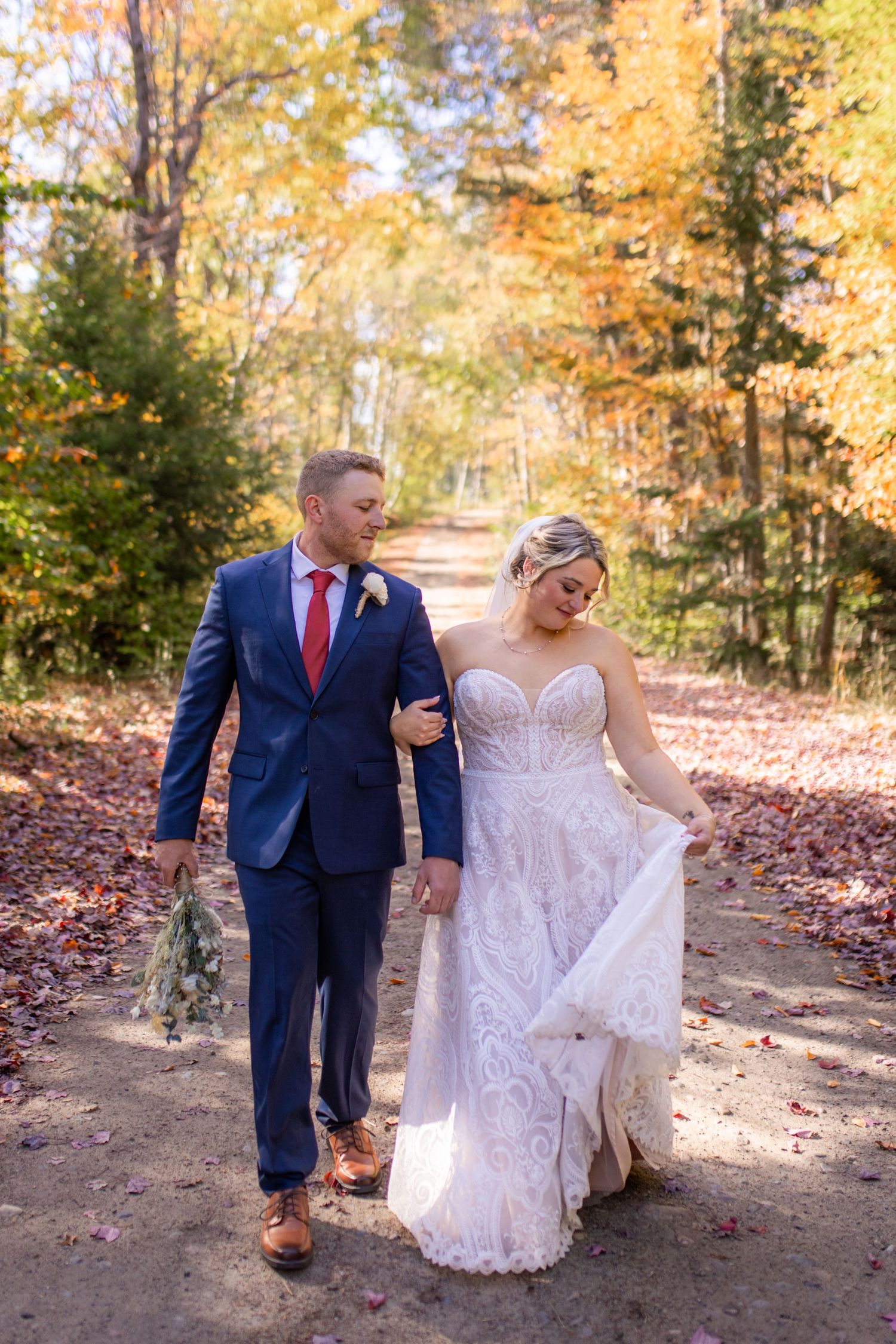 A couple in wedding attire walks together on a leaf-covered path surrounded by autumn trees with golden foliage.