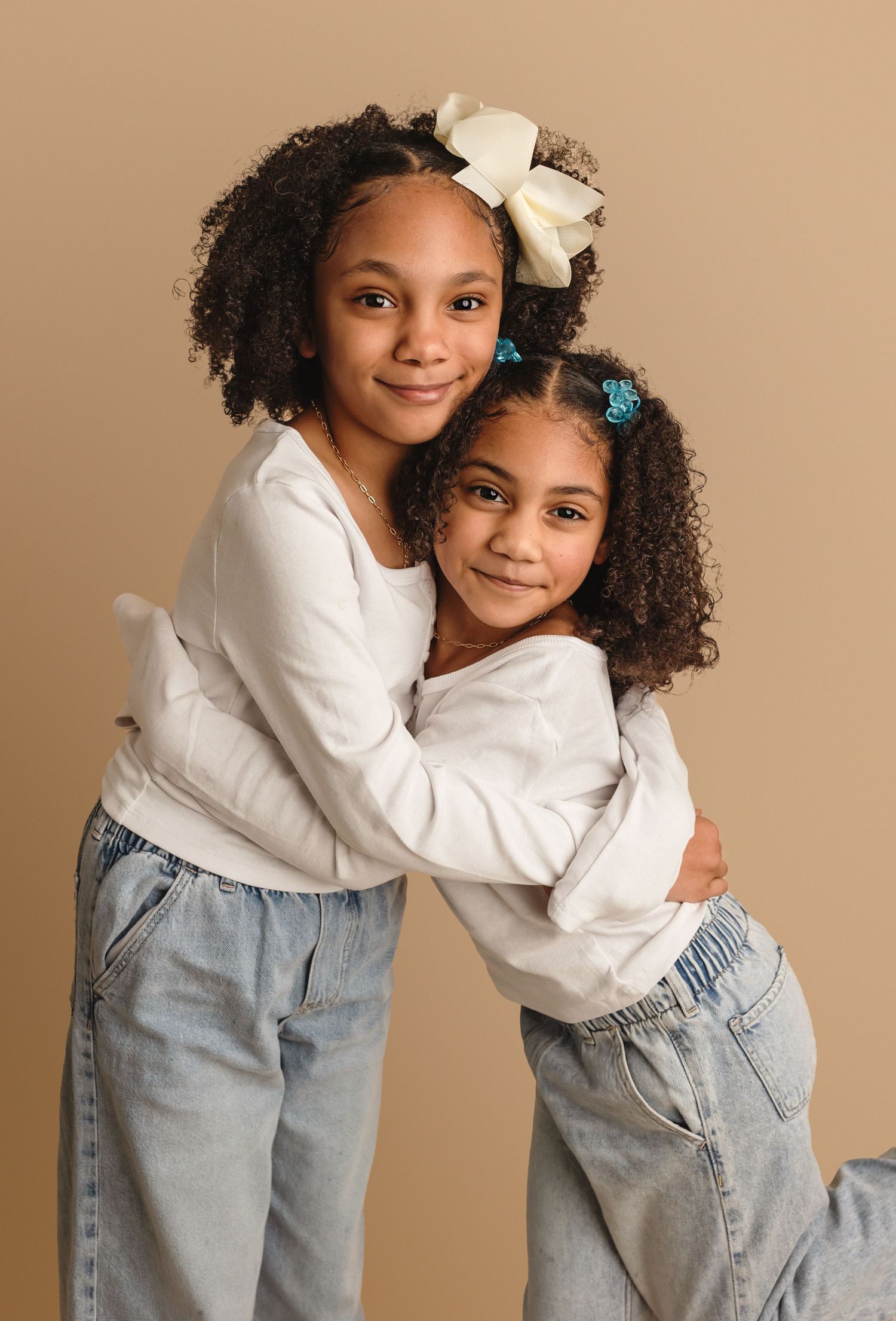 Young sisters in white shirts and jeans hug at Somerset Portraits newborn and baby photography studio for their family photography session.