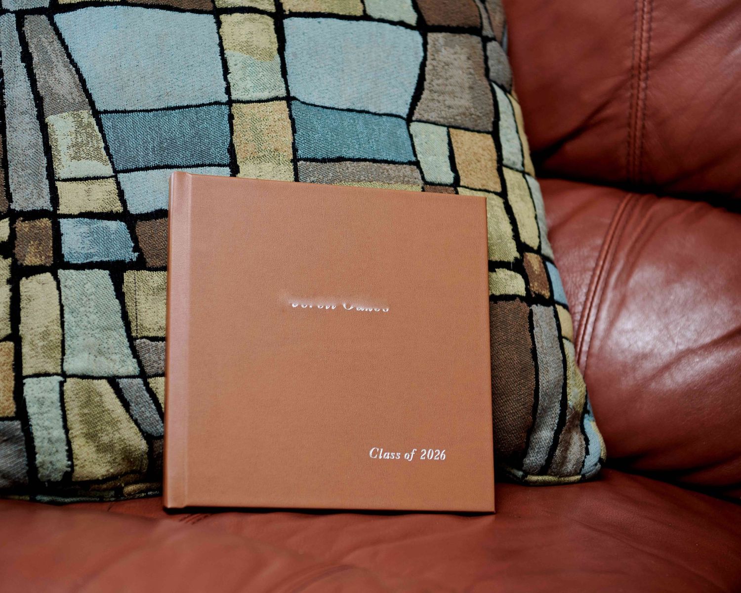 High school senior "Memory" album in honey leatherette, resting against a blue checkered pillow on brown leather couch.