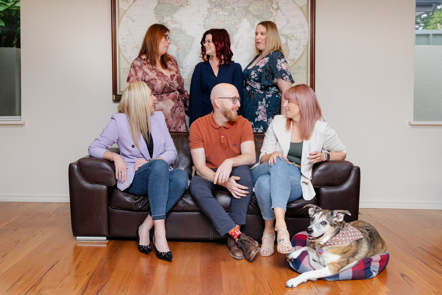 A group sits together on a brown leather couch with a husky mix dog in a bright interior setting.