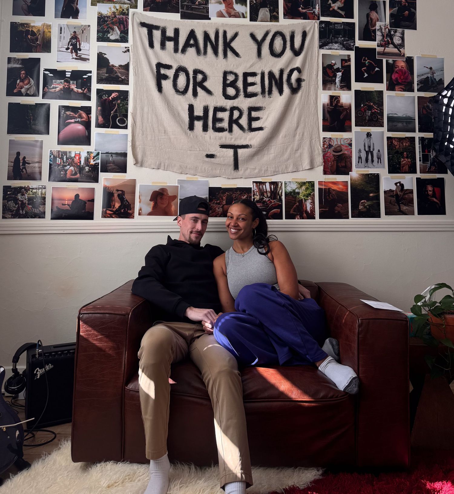 Two people sit together on a brown leather couch in a room with photo-covered walls and a 'Thank You' banner.