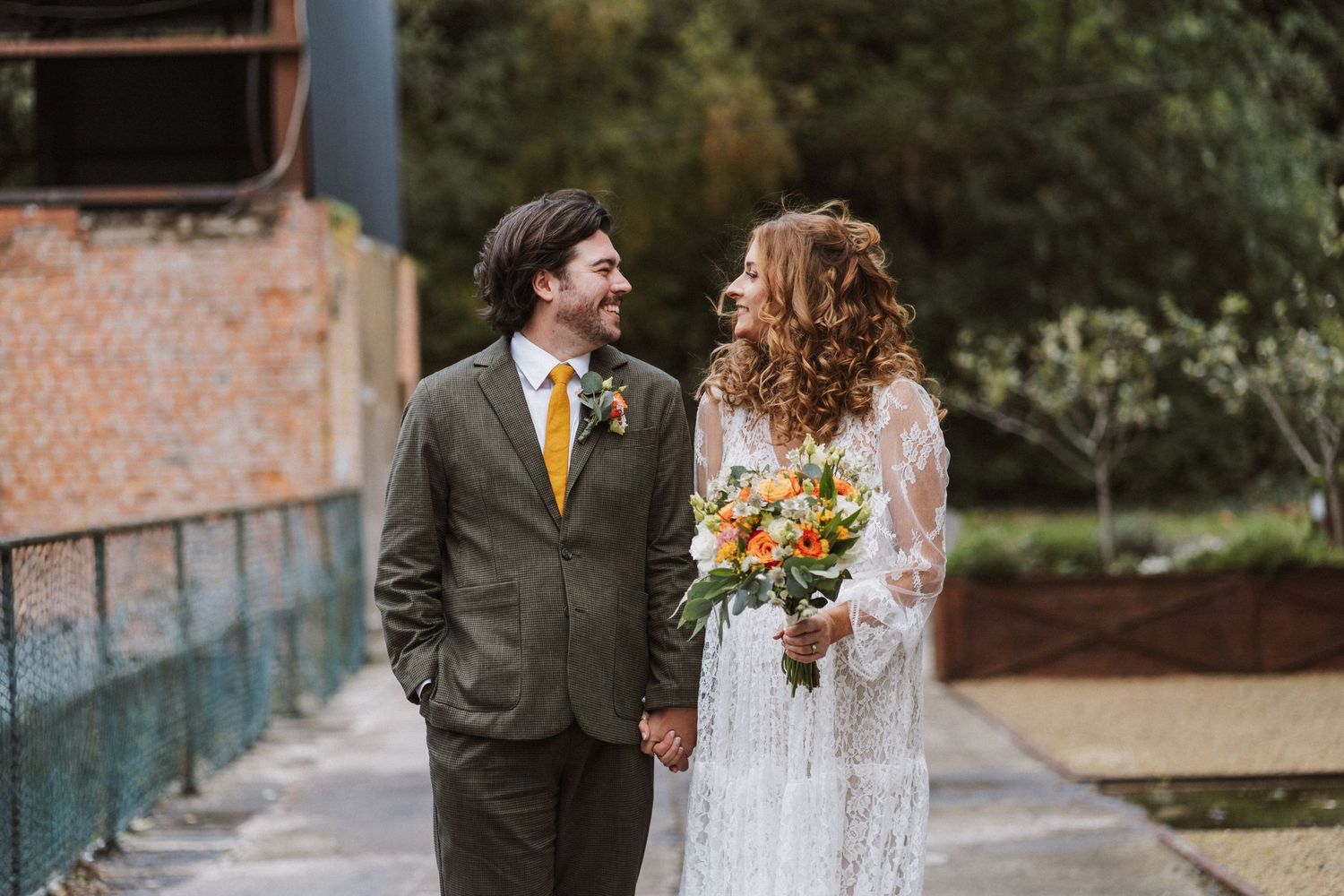 A couple in wedding attire walks together through a garden path surrounded by lush greenery.