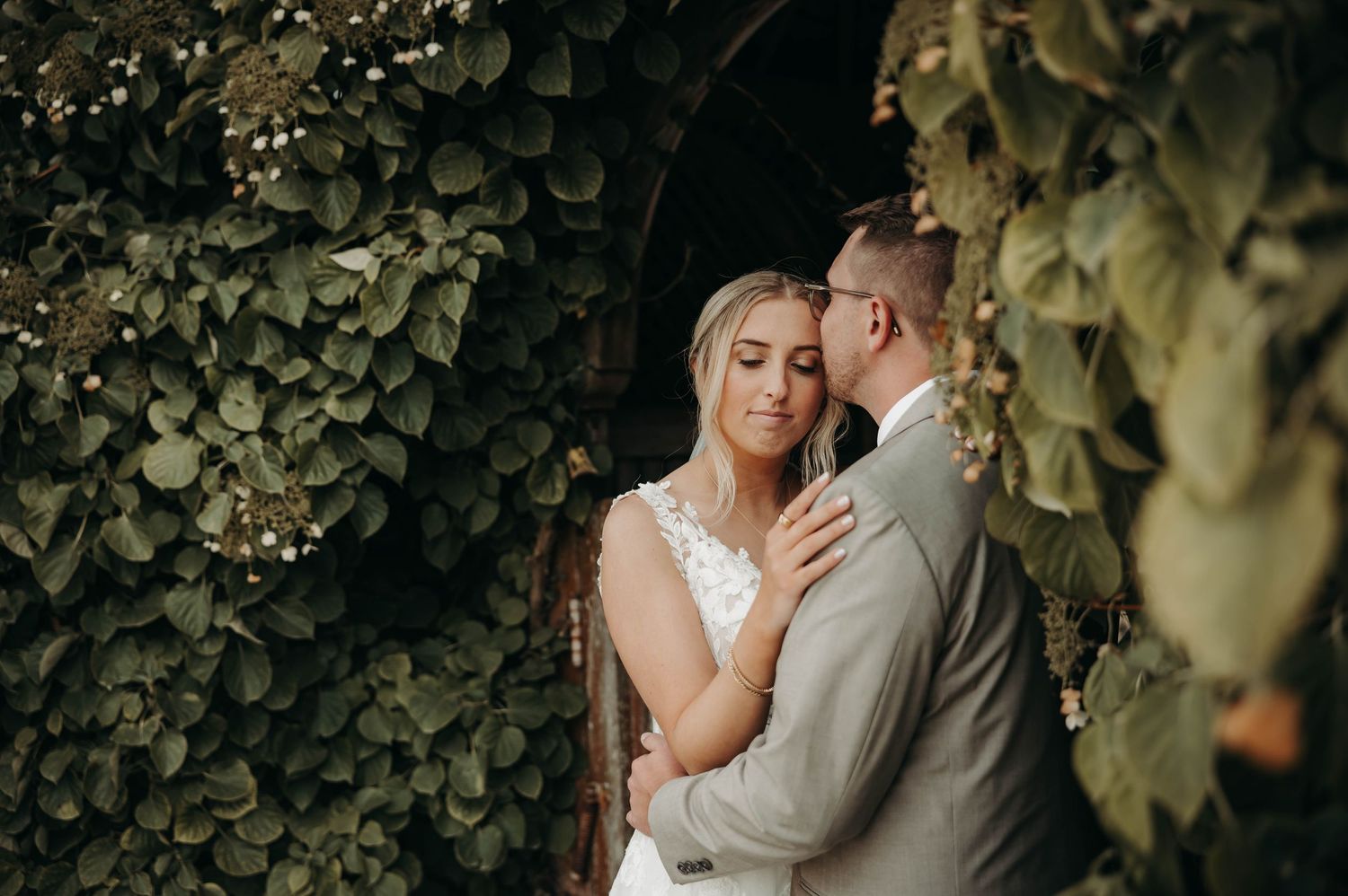 Bride and groom sharing tender embrace beneath lush green ivy-covered archway at outdoor wedding.