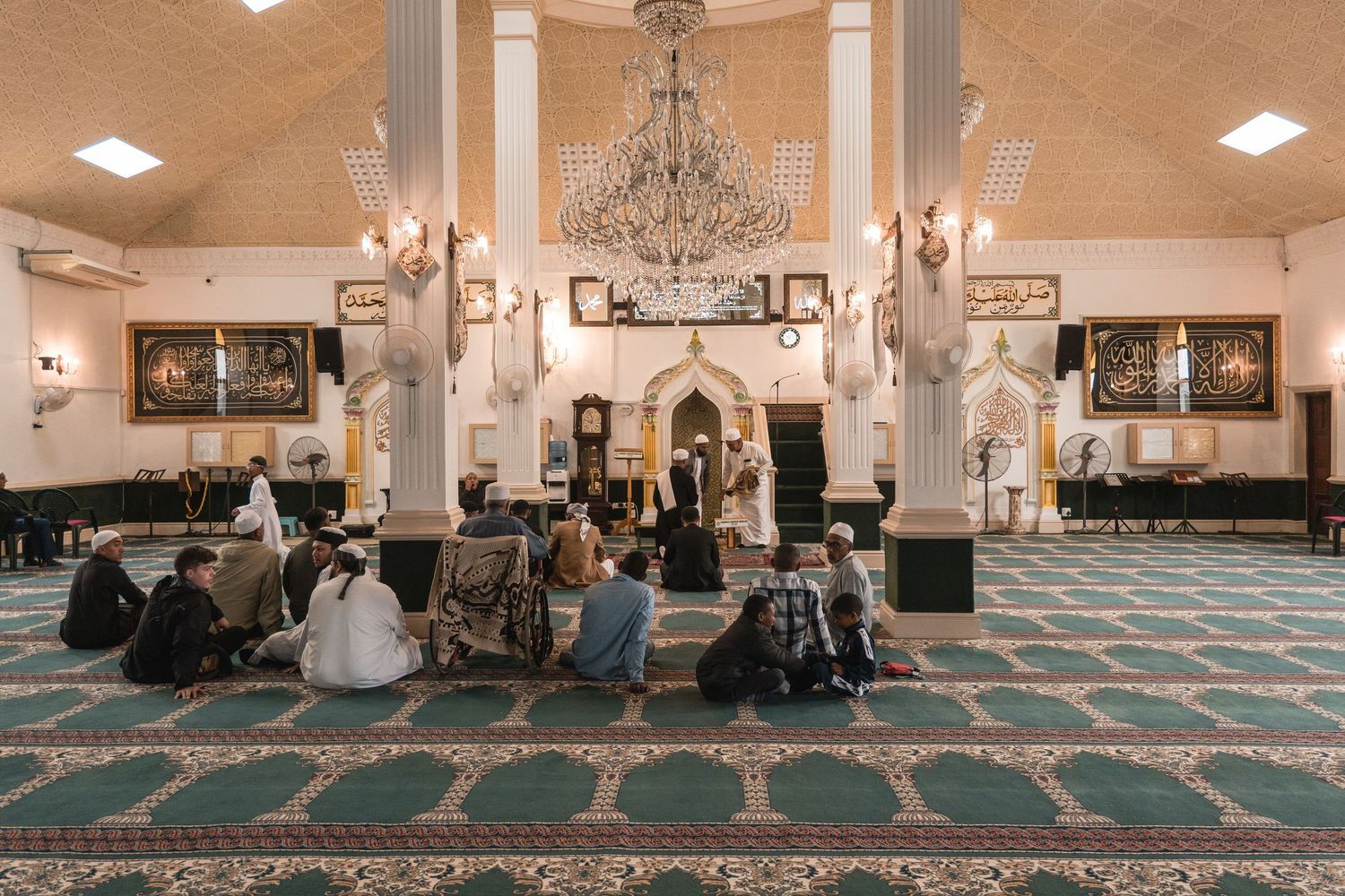 Interior of mosque with chandelier and people gathered on ornate turquoise carpet.
