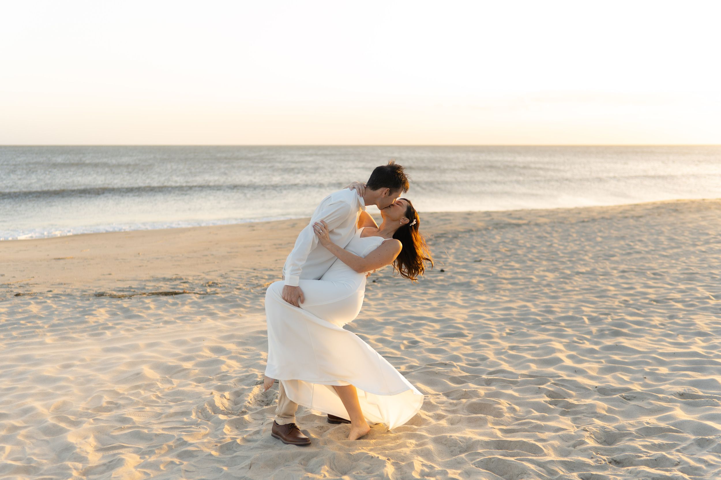 Couple in white attire shares romantic moments during sunset beach photoshoot.