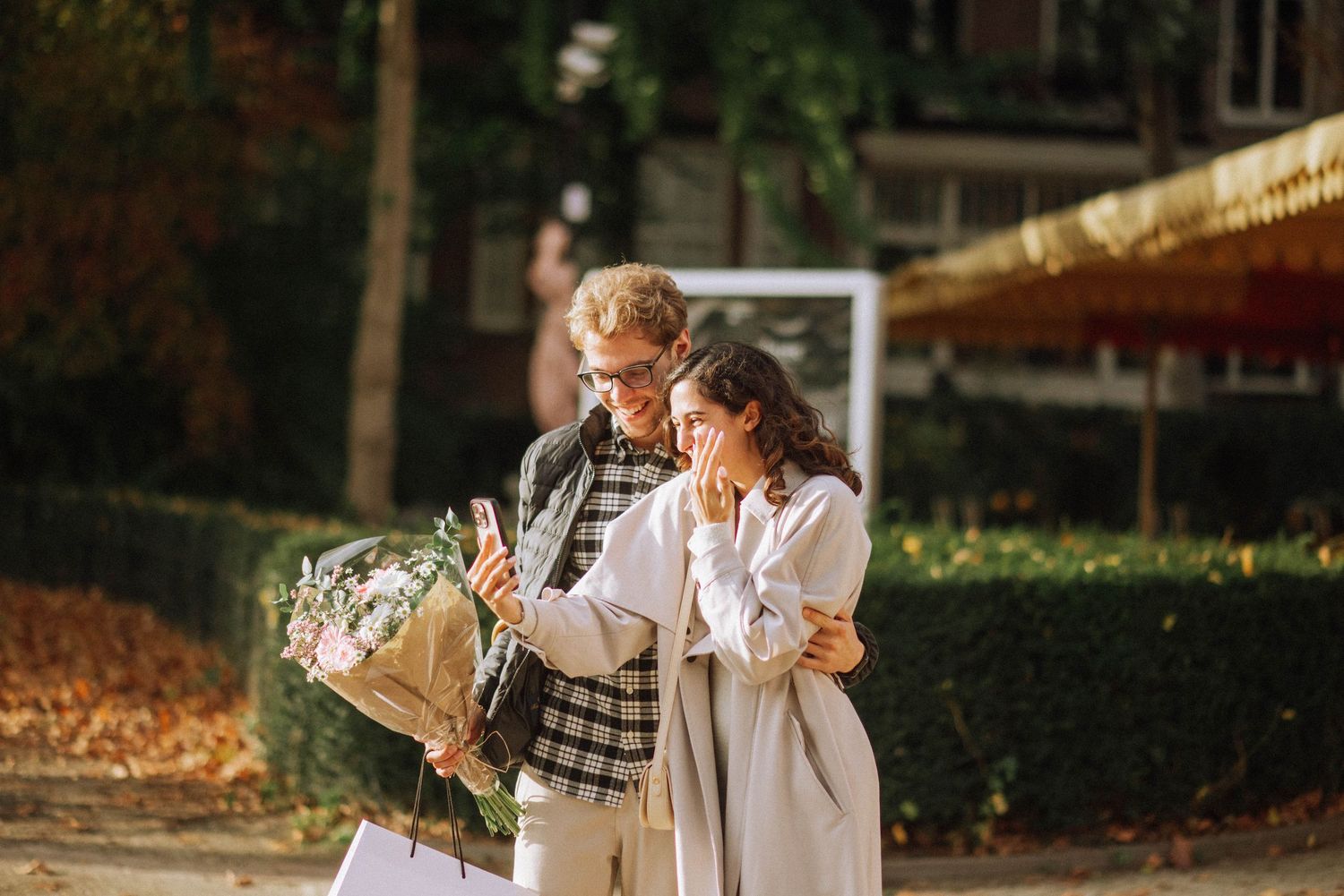 Engaged couple taking a selfie in an Amsterdam Vondelpark with a bouquet of flowers, celebrating their proposal in autumn.