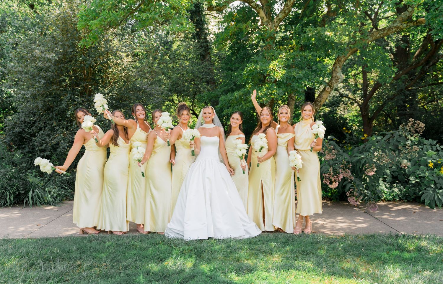 A wedding party in light yellow bridesmaid dresses poses together in a lush garden setting.