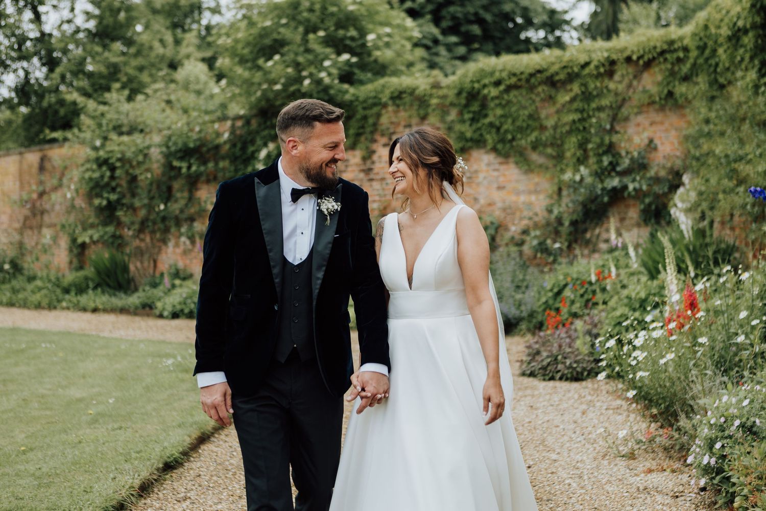 A couple in formal wedding attire walks together in a garden path surrounded by lush greenery and stone walls.
