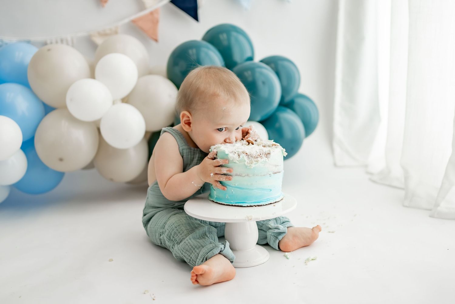 Baby in pastel green and blue clothing smashes cake at first birthday photoshoot decorated with balloons.