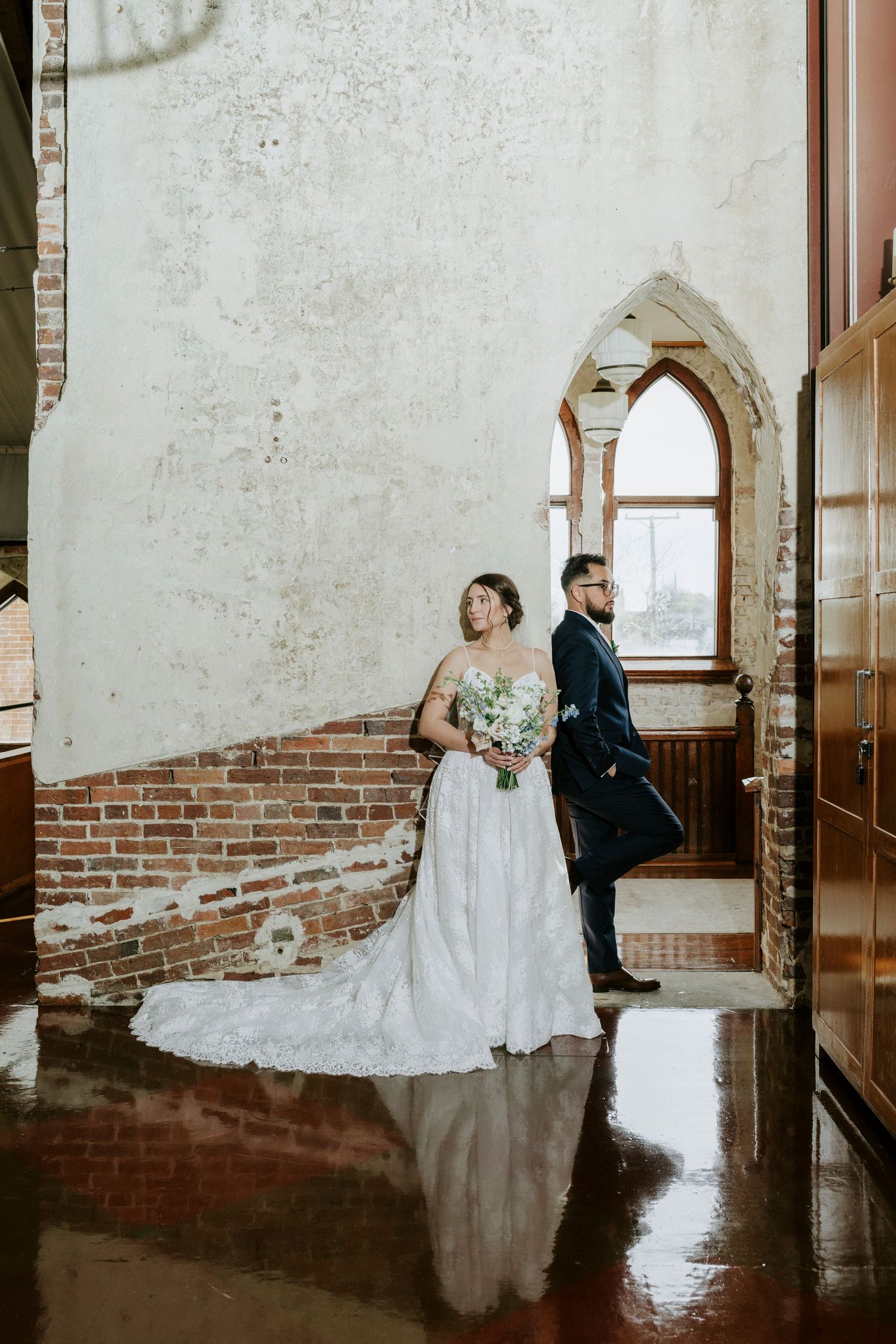 Wedding couple poses in a rustic brick venue with elegant arched windows and reflective floors.