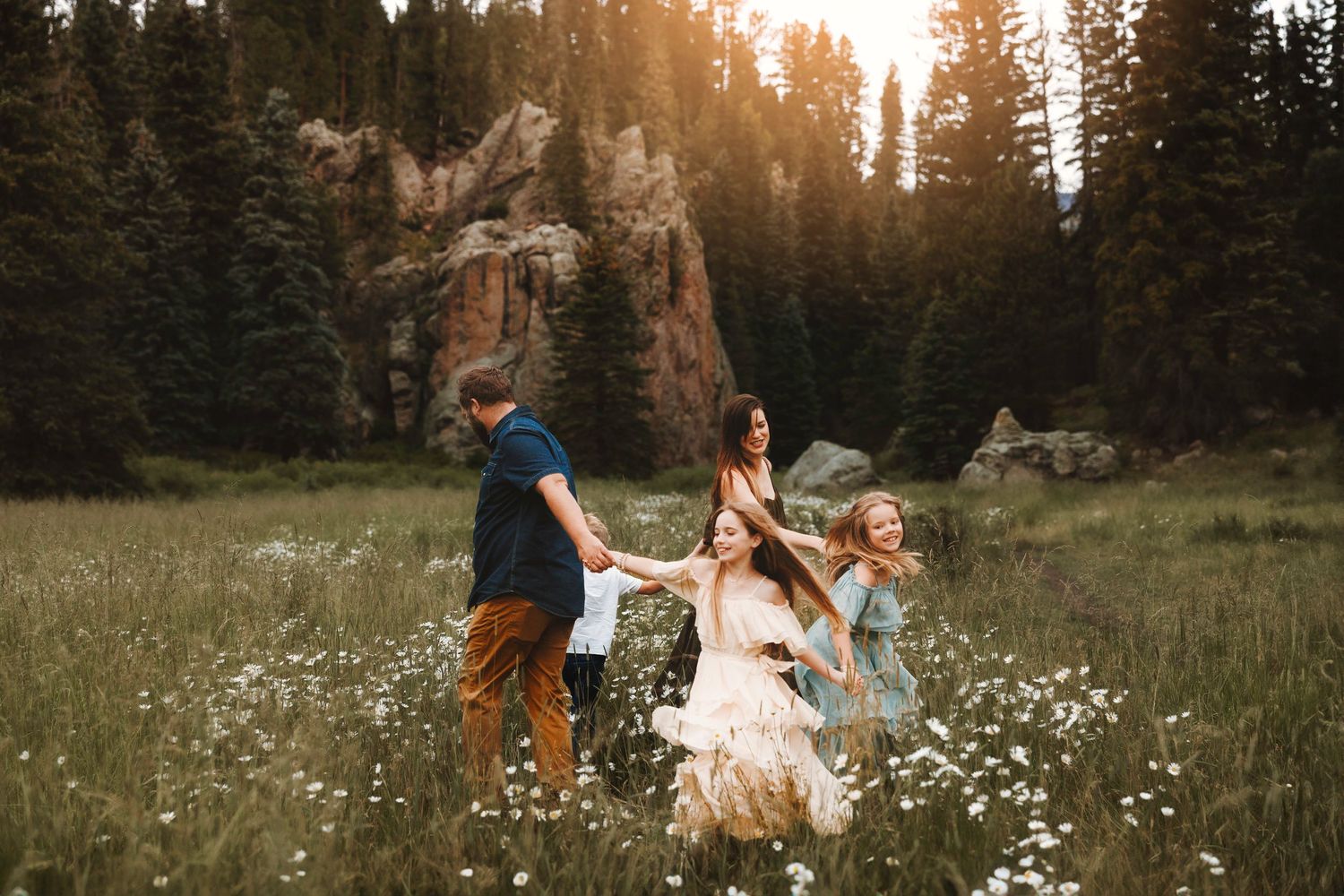 Family running through a wildflower meadow with pine trees and rocky cliffs at sunset.