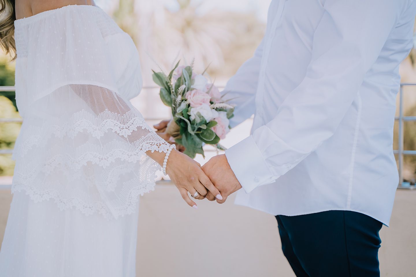 Close up of bride and groom holding hands with white dress and green floral bouquet at wedding ceremony.
