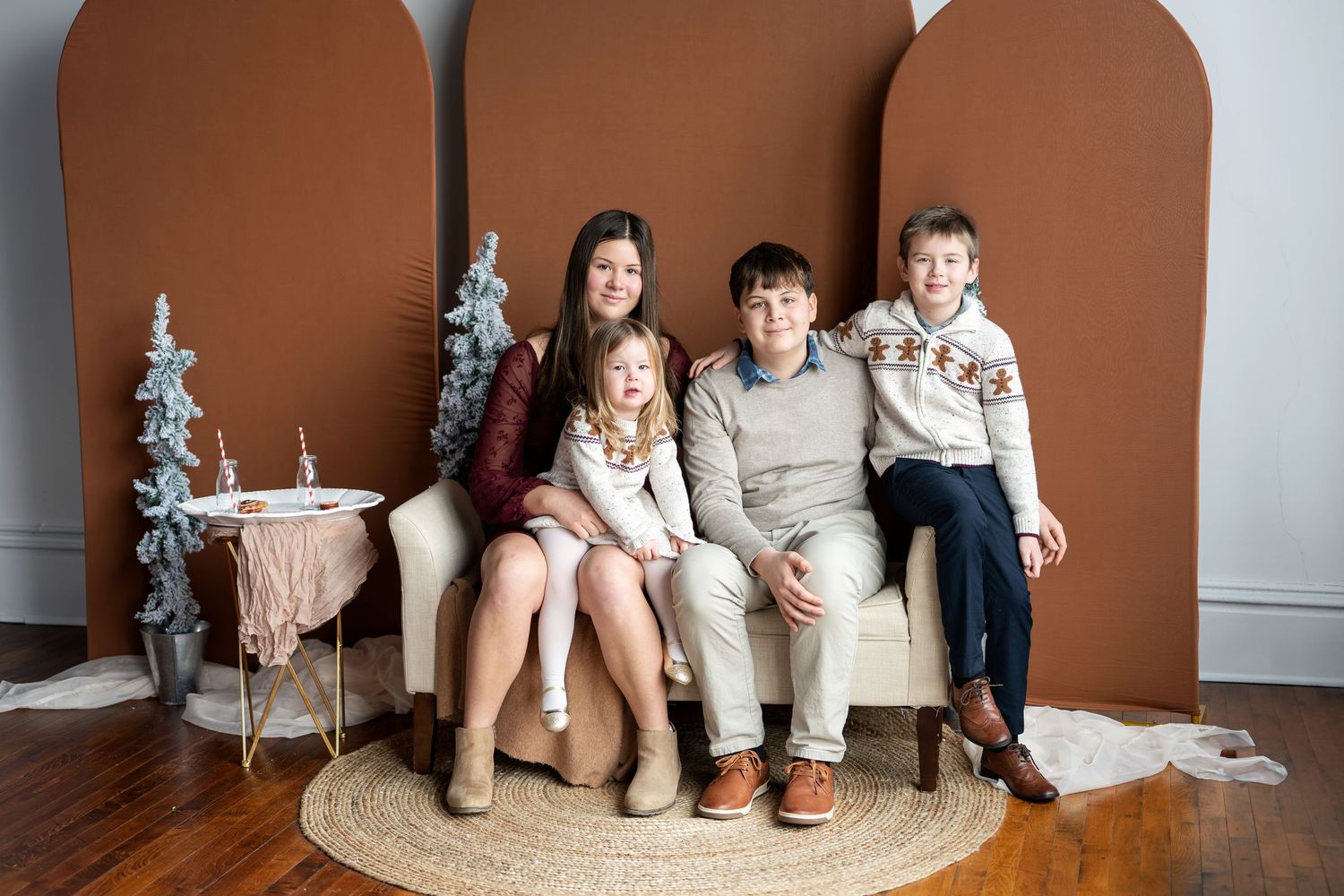Four siblings posing together on a white couch with arched brown backdrops and Christmas trees.