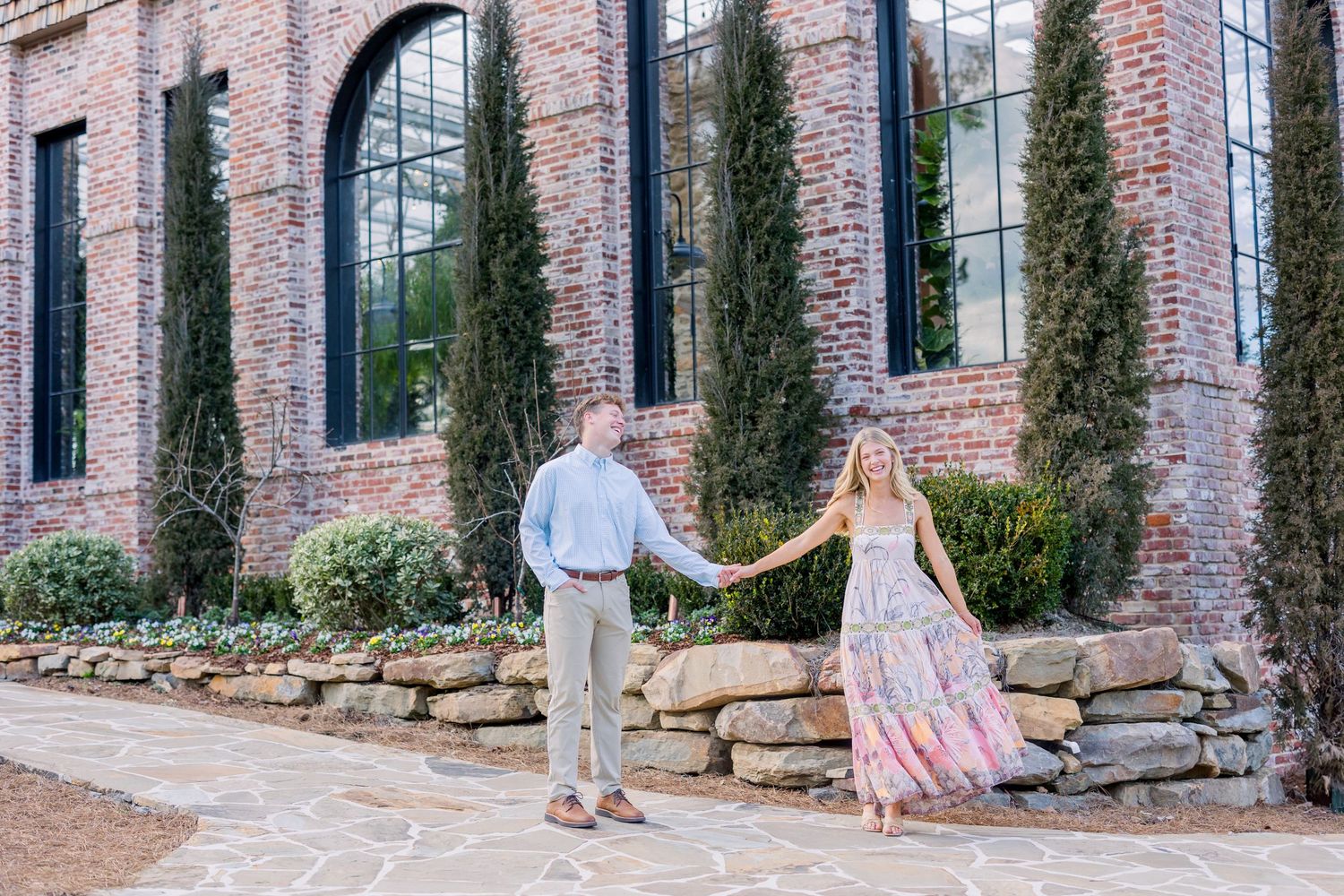 A couple walks along a brick building exterior with tall cypress trees and stone retaining wall.
