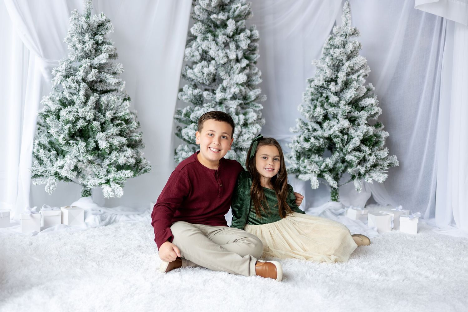 Two children in holiday outfits sitting together on white fluffy floor near snowy Christmas trees.
