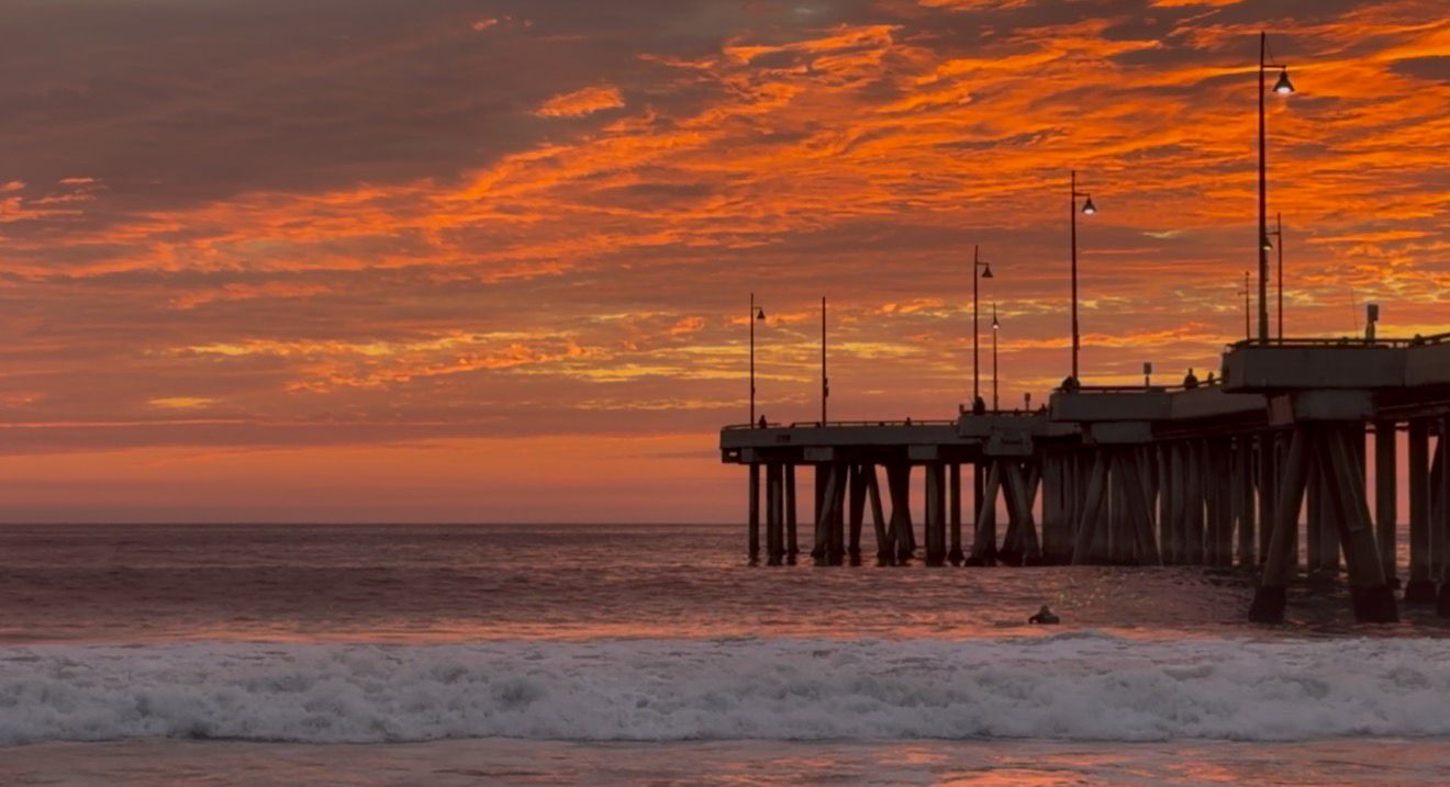 Dramatic orange and red sunset sky over a pier silhouetted against the Pacific Ocean waves.