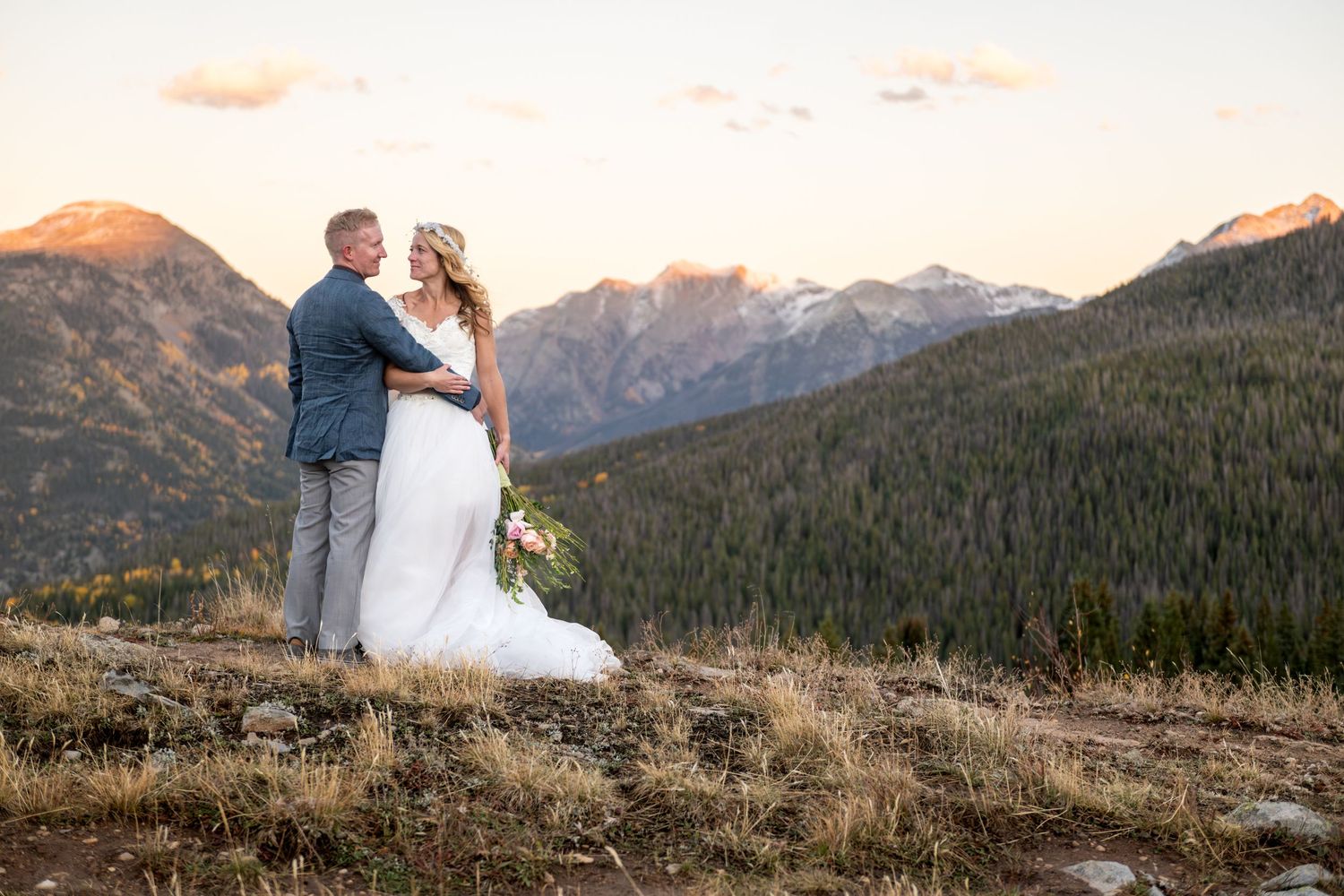 A bride and groom embrace on a mountain overlook at sunset with rugged mountain peaks and pine forests in the background.