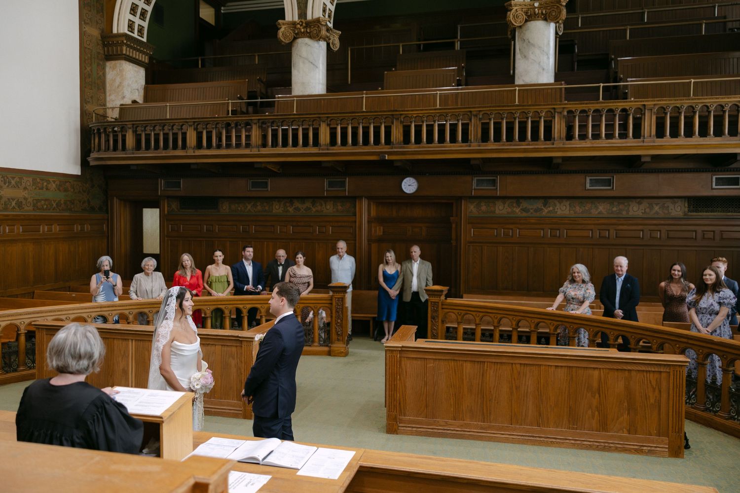 Intimate civil wedding ceremony in historic wood-paneled courtroom with small group of seated guests.