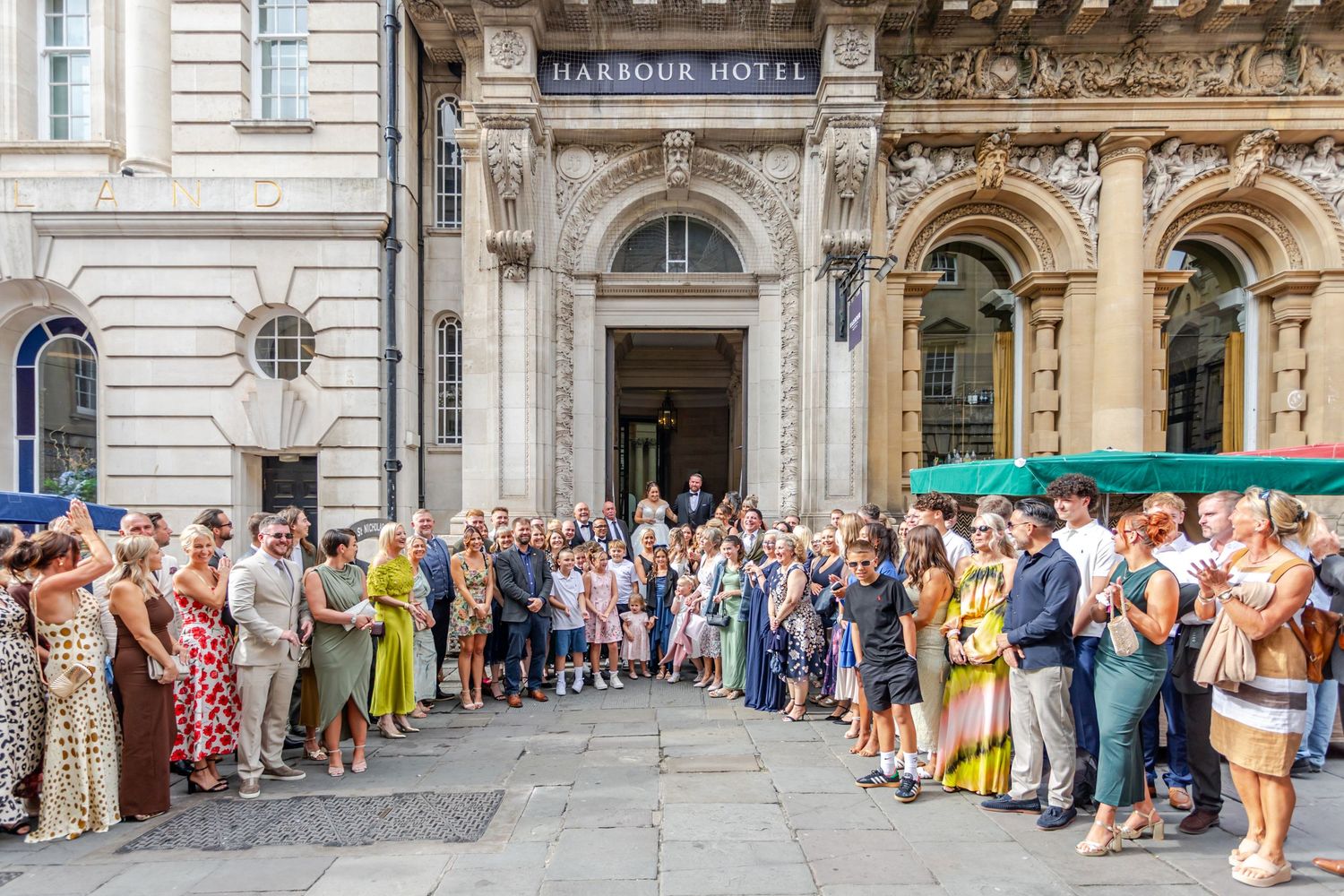 Large wedding party gathered outside the elegant Harbour Hotel entrance on a sunny day.